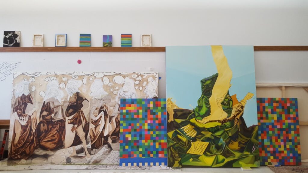 Studio wall hung with large figurative and abstract paintings and small grid works on a shelf.